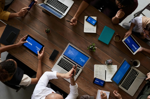 A group of people sitting around a wooden table, each engaged with laptops, tablets, and smartphones displaying blue screens with text. The setting suggests a collaborative work or