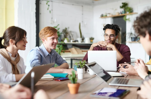 A group of people sitting around a wooden table in a casual office setting, engaged in a discussion. Laptops and notebooks are on the table, and the background features plants and s